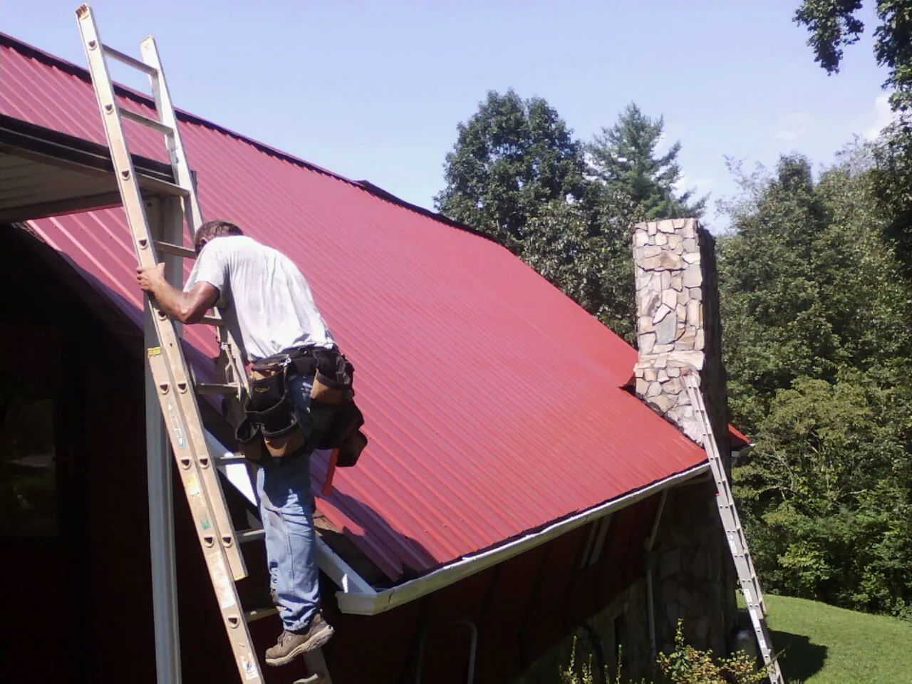 Roofer on a ladder beside a red metal roof and stone chimney — Soffit & Fascia Repair by Roofing Brothers in Corsicana